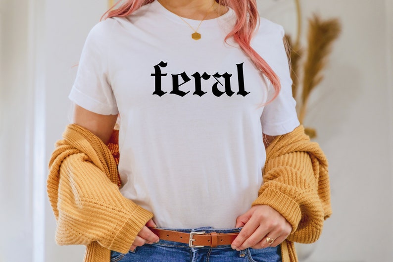 Puede incluir: Una camiseta blanca con la palabra "feral" impresa en letras g&oacute;ticas negras. La camiseta la lleva puesta una persona con el pelo rosa y un c&aacute;rdigan amarillo.