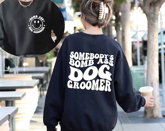 May include: Black sweatshirt with white text that reads "Somebody's Bomb Ass Dog Groomer" in a retro font. The sweatshirt has a crew neck and long sleeves.