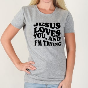 Pu&ograve; includere: Una maglietta grigia con la scritta nera "JESUS LOVES YOU, AND I'M TRYING". La maglietta ha maniche corte e girocollo. La persona che indossa la maglietta ha la mano sul fianco.