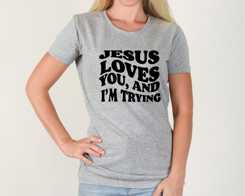 Pu&ograve; includere: Una maglietta grigia con la scritta nera "JESUS LOVES YOU, AND I'M TRYING". La maglietta ha maniche corte e girocollo. La persona che indossa la maglietta ha la mano sul fianco.