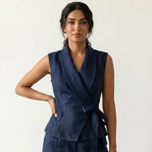 May include: A sleeveless navy blue wrap vest featuring a wide shawl collar and a side tie closure, paired with matching wide-leg trousers. The set is crafted from a textured fabric with a visible weave, creating a cohesive and structured appearance.