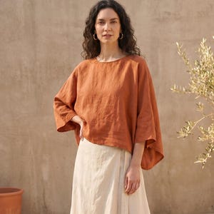 May include: A burnt orange, loose-fitting top with three-quarter sleeves and a cream-coloured wrap skirt. The outfit is made of a lightweight fabric. The model is wearing gold hoop earrings. The background includes a terracotta pot and a small tree.