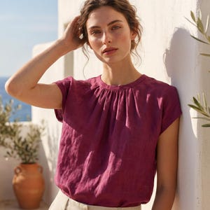 May include: A deep magenta short-sleeved blouse with a round neckline and subtle vertical pleats at the collar, paired with light beige trousers featuring a structured waistband and front pleats.