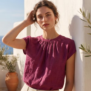 May include: A magenta-coloured short-sleeved top with a gathered neckline, worn with light beige trousers. The background includes a white wall, olive trees in terracotta pots, and a view of the sea.