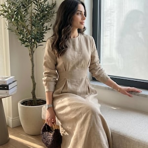 Puede incluir: Un vestido largo de color beige con mangas largas, cuello redondo y una cintura entallada con pliegues sutiles. La prenda está confeccionada en un tejido texturizado e incluye mangas largas con puños, lo que proporciona una silueta estructurada.