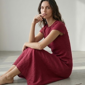 May include: A burgundy-colored dress with short sleeves. The dress has a round neckline and a flowing skirt. The model is sitting on a light gray surface. The dress appears to be made of linen or a similar fabric.