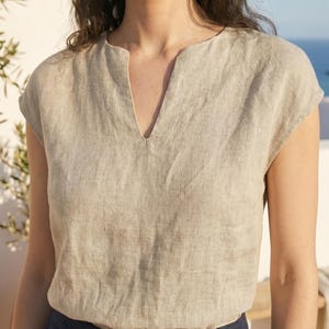 May include: A beige linen top with a V-neckline and cap sleeves. The top is made of a textured fabric and is paired with navy blue bottoms. The model is wearing gold hoop earrings. The background features a white wall and a glimpse of the ocean.