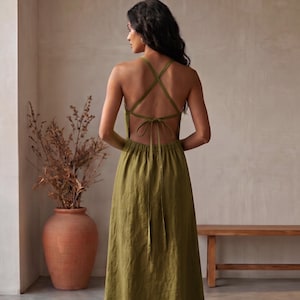 May include: An olive green midi dress featuring a backless design with thin crisscross straps that tie at the waist. The garment has a gathered elasticized waistband and a flowing skirt that falls to the mid-calf, crafted from a textured fabric.