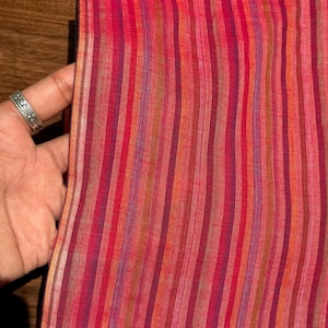 Może przedstawiać: Zbliżenie tkaniny z pionowymi paskami w odcieniach czerwieni, różu, pomarańczy i fioletu. Tkanina trzymana jest przez dłoń z srebrnym pierścionkiem. Paski różnią się szerokością i intensywnością koloru, tworząc teksturowany wygląd.