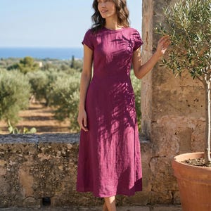 May include: A magenta-colored, short-sleeved dress with a round neckline. The dress is knee-length and made of a textured fabric. The background features a stone wall, olive trees, and a view of the sea.