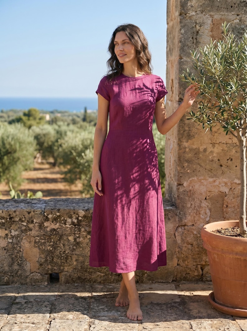 May include: A magenta-colored, short-sleeved dress with a round neckline. The dress is knee-length and made of a textured fabric. The background features a stone wall, olive trees, and a view of the sea.