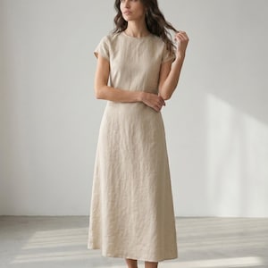 May include: A beige linen dress with a round neckline and short sleeves. The dress has a midi length, reaching the mid-calf. The fabric appears to be lightweight and flowing, suitable for warm weather. The dress is a solid color with a simple, elegant design.