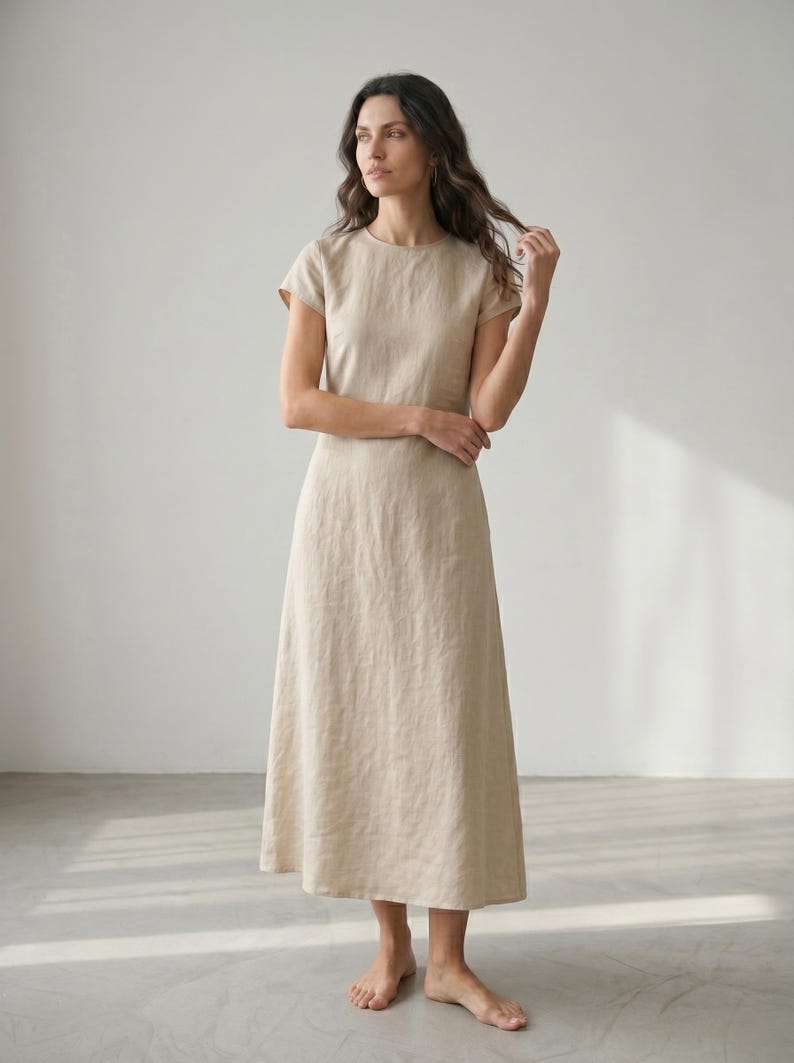 May include: A beige linen dress with a round neckline and short sleeves. The dress has a midi length, reaching the mid-calf. The fabric appears to be lightweight and flowing, suitable for warm weather. The dress is a solid color with a simple, elegant design.