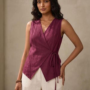 A woman wearing a purple sleeveless top with a tie at the waist, posing against a beige background.