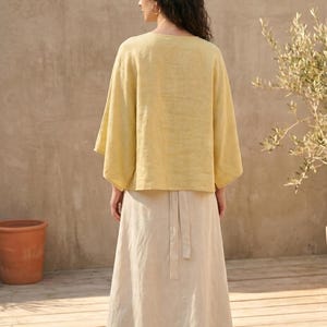 May include: A woman wearing a yellow linen top with wide sleeves and a long, cream-colored skirt. The outfit is simple and elegant, suitable for warm weather. The background features a neutral-toned wall and wooden flooring, with a potted plant on the left.