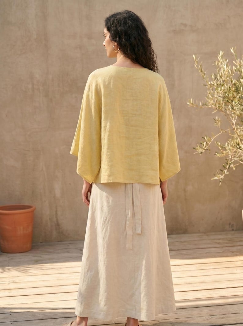 May include: A woman wearing a yellow linen top with wide sleeves and a long, cream-colored skirt. The outfit is simple and elegant, suitable for warm weather. The background features a neutral-toned wall and wooden flooring, with a potted plant on the left.