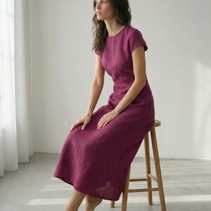 May include: A magenta-colored dress with short sleeves and a round neckline. The dress is knee-length and appears to be made of linen. The person is seated on a wooden stool in a room with natural light.