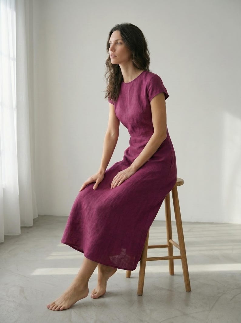 May include: A magenta-colored dress with short sleeves and a round neckline. The dress is knee-length and appears to be made of linen. The person is seated on a wooden stool in a room with natural light.