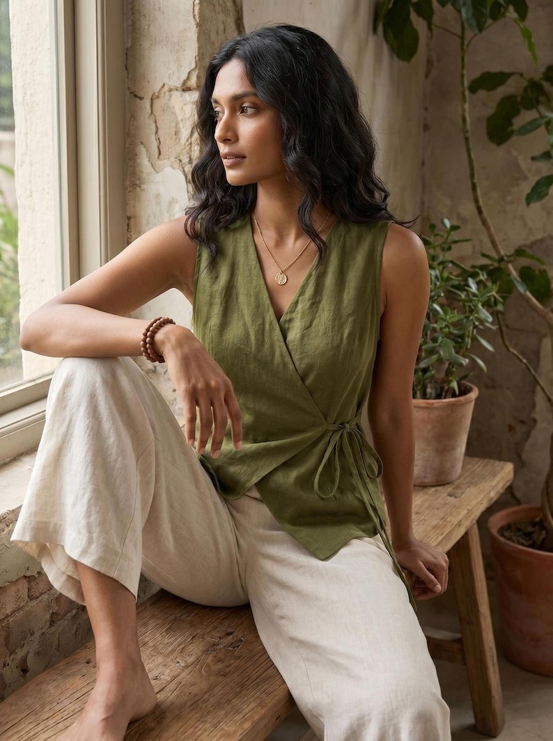 A woman with long dark hair is sitting on a wooden bench, wearing a green sleeveless top and white pants. She is looking out a window, with potted plants visible in the background.