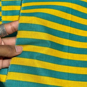 Op de afbeelding: Close-up van een stof met horizontale strepen in afwisselende kleuren van teal en geel. De stof wordt vastgehouden, waardoor de rafelige rand zichtbaar wordt. De strepen zijn gelijkmatig verdeeld en cre&euml;ren een gedurfd, grafisch patroon.