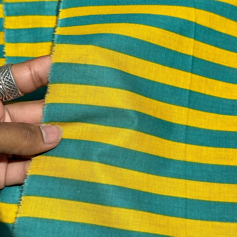 Op de afbeelding: Close-up van een stof met horizontale strepen in afwisselende kleuren van teal en geel. De stof wordt vastgehouden, waardoor de rafelige rand zichtbaar wordt. De strepen zijn gelijkmatig verdeeld en cre&euml;ren een gedurfd, grafisch patroon.