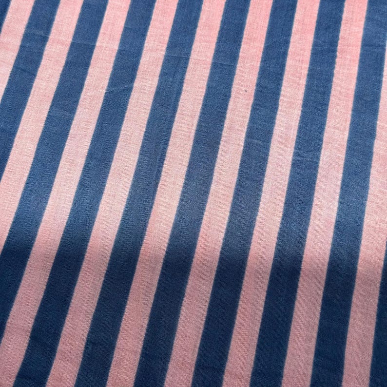 Op de afbeelding: Close-up van een stof met een herhalend patroon van blauwe en roze strepen. De strepen zijn diagonaal en gelijkmatig verdeeld, waardoor een visueel aantrekkelijk ontwerp ontstaat. De stof lijkt zacht en licht van gewicht.