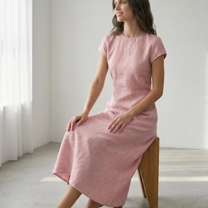 May include: A light pink linen dress with short sleeves and a round neckline. The dress is knee-length and has a simple, elegant design. The model is sitting on a wooden stool in a well-lit room.
