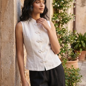 May include: A sleeveless, off-white linen top with a mandarin collar and decorative floral-shaped buttons. The top is paired with black linen pants. The model is leaning against a stone wall, with potted plants in the background.