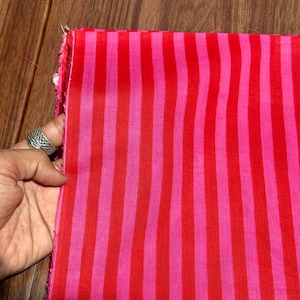 Op de afbeelding: Een stuk stof met verticale strepen in afwisselende tinten rood en roze. De stof wordt vastgehouden door een hand met een zilveren ring. De randen van de stof zijn onafgewerkt.