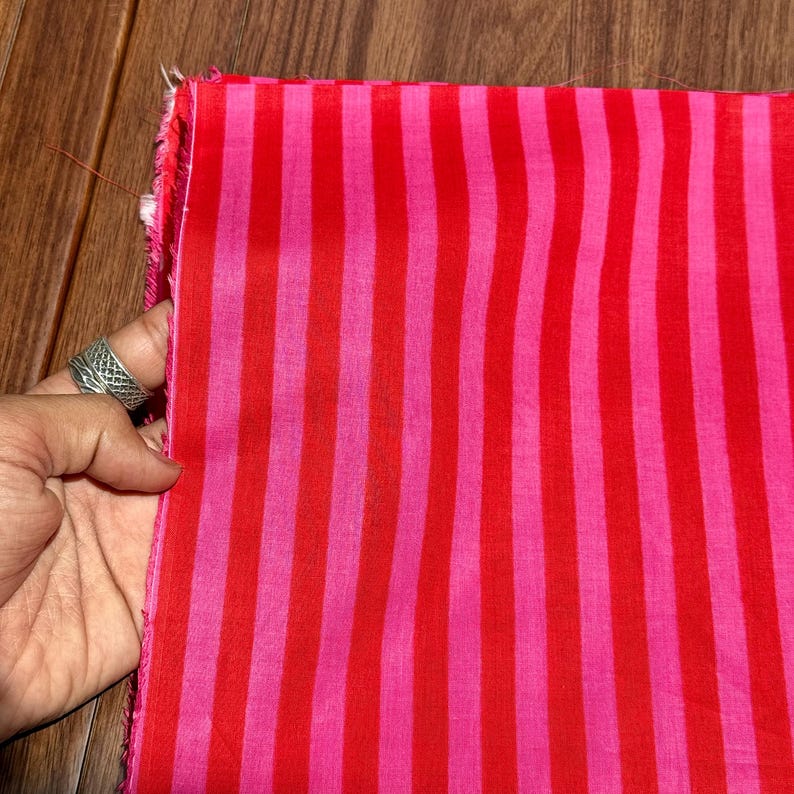 Op de afbeelding: Een stuk stof met verticale strepen in afwisselende tinten rood en roze. De stof wordt vastgehouden door een hand met een zilveren ring. De randen van de stof zijn onafgewerkt.