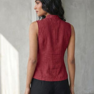 May include: A sleeveless, burgundy-colored top with a high collar. The top is made of a textured fabric and has a fitted silhouette. The model is also wearing black pants. The background is a plain, light gray wall.