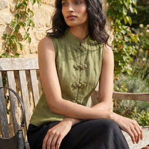May include: Olive green sleeveless top with a mandarin collar and decorative frog closures. The top is paired with black pants. A black handbag rests on a wooden bench in an outdoor setting.