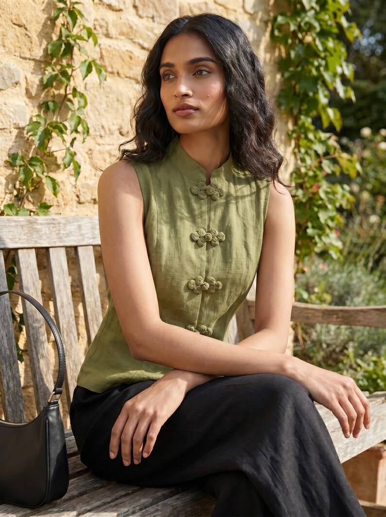 May include: Olive green sleeveless top with a mandarin collar and decorative frog closures. The top is paired with black pants. A black handbag rests on a wooden bench in an outdoor setting.