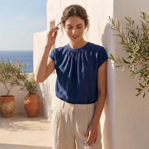 Peut inclure: Un haut en lin bleu marine avec un col fronc&eacute; et des manches courtes. Le haut est associ&eacute; &agrave; un pantalon en lin beige. L'image est prise &agrave; l'ext&eacute;rieur avec des oliviers dans des pots en terre cuite et un mur blanc.