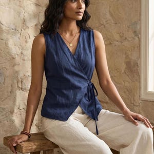 A woman with long dark hair is sitting on a wooden stool, wearing a blue sleeveless top and white pants. She is looking off to the side, with a neutral expression on her face.