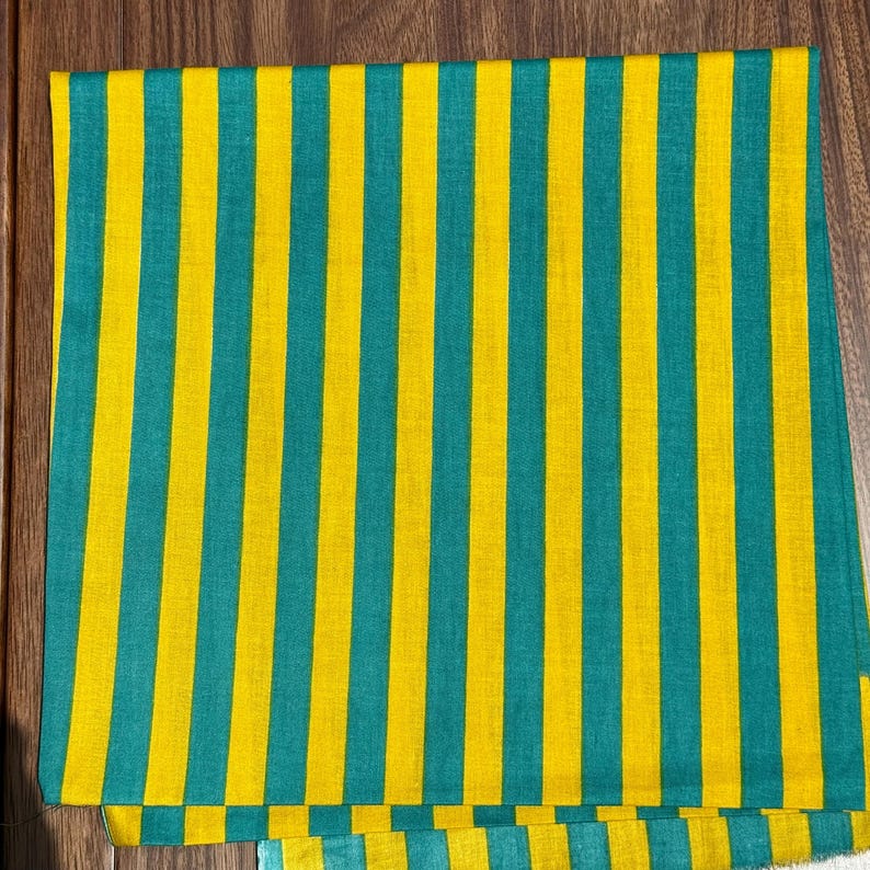 Op de afbeelding: Een gevouwen textiel met een herhalend patroon van verticale strepen. De strepen wisselen af tussen een helder geel en een blauwgroene kleur. Het textiel ligt op een houten oppervlak.