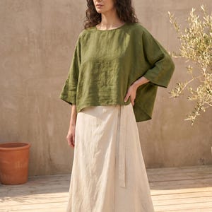 May include: An olive green linen top with wide sleeves and a cream-colored wrap skirt. The skirt has a matching fabric belt. The outfit is casual and comfortable, suitable for warm weather. The model is standing outdoors.