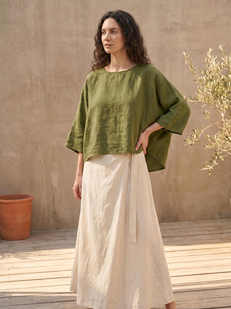 May include: An olive green linen top with wide sleeves and a cream-colored wrap skirt. The skirt has a matching fabric belt. The outfit is casual and comfortable, suitable for warm weather. The model is standing outdoors.
