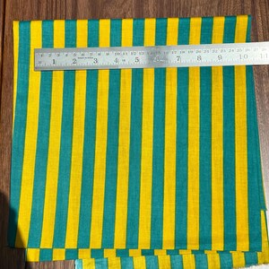 Op de afbeelding: Een stof met verticale strepen in geel en teal. Een metalen liniaal is over de stof geplaatst, met metingen in centimeters. Het patroon van de stof is consistent, met gelijkmatig verdeelde strepen.