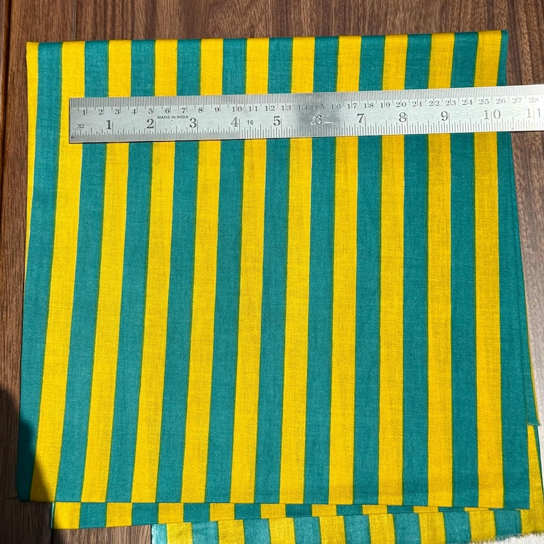 Op de afbeelding: Een stof met verticale strepen in geel en teal. Een metalen liniaal is over de stof geplaatst, met metingen in centimeters. Het patroon van de stof is consistent, met gelijkmatig verdeelde strepen.