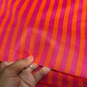 Op de afbeelding: Close-up van een stof met verticale strepen in afwisselende tinten roze en oranje. De stof wordt aan de rand vastgehouden, waardoor de textuur en kleurvariaties zichtbaar worden. De strepen zijn gelijkmatig verdeeld.