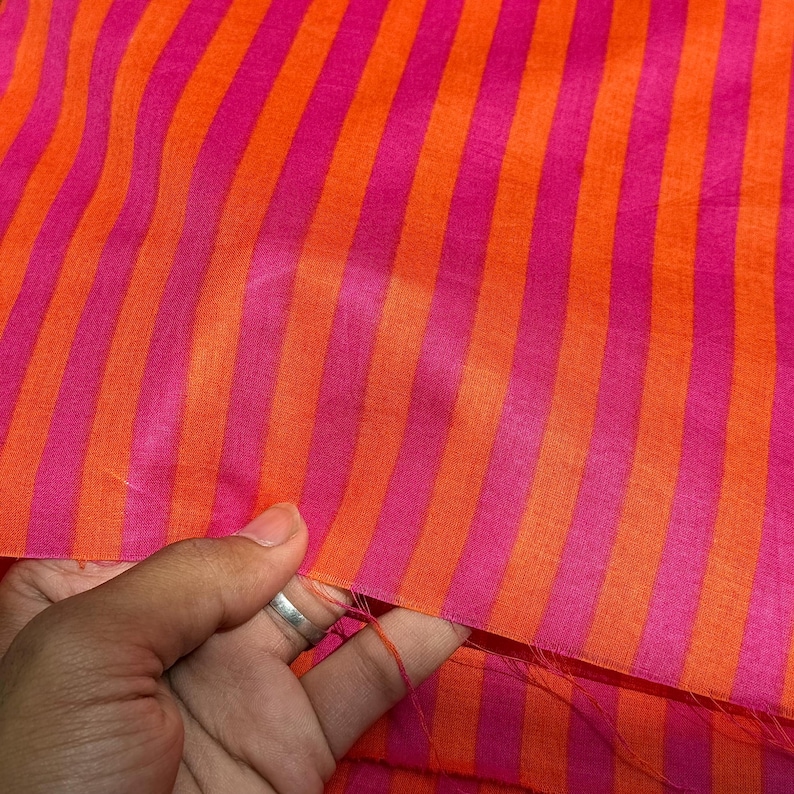 Op de afbeelding: Close-up van een stof met verticale strepen in afwisselende tinten roze en oranje. De stof wordt aan de rand vastgehouden, waardoor de textuur en kleurvariaties zichtbaar worden. De strepen zijn gelijkmatig verdeeld.