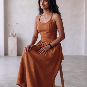 May include: A rust-colored midi dress featuring a scoop neckline, thin shoulder straps, and a fitted bodice that transitions into a gathered, flowing skirt. The garment is constructed from a solid-colored fabric with a visible, textured weave.