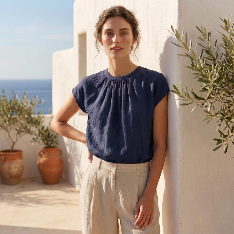Peut inclure: Un haut en lin bleu marine avec un col fronc&eacute; et des manches courtes. Il est associ&eacute; &agrave; un pantalon en lin beige. Le cadre est en ext&eacute;rieur, avec un mur blanc et des oliviers dans des pots en terre cuite. La mer est visible en arri&egrave;re-plan.