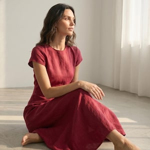 May include: A burgundy-colored linen dress with short sleeves. The dress has a round neckline and a flowing skirt. The person is sitting on the floor with their legs crossed. The dress appears to be made of a lightweight fabric.