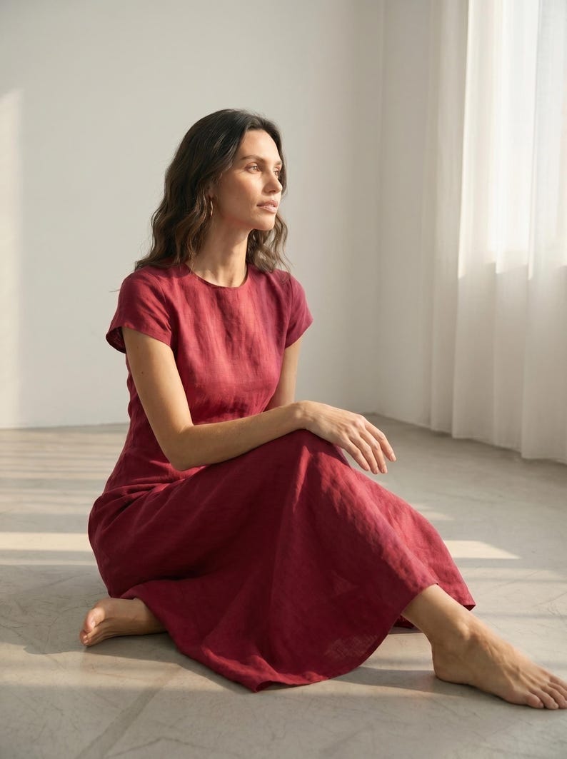 May include: A burgundy-colored linen dress with short sleeves. The dress has a round neckline and a flowing skirt. The person is sitting on the floor with their legs crossed. The dress appears to be made of a lightweight fabric.