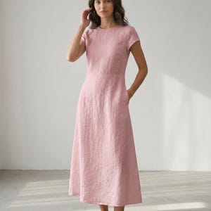 May include: A light pink midi dress with short sleeves and a round neckline. The dress has a fitted bodice and a flared skirt, with pockets on each side. The model is wearing the dress and standing in a room with natural light.