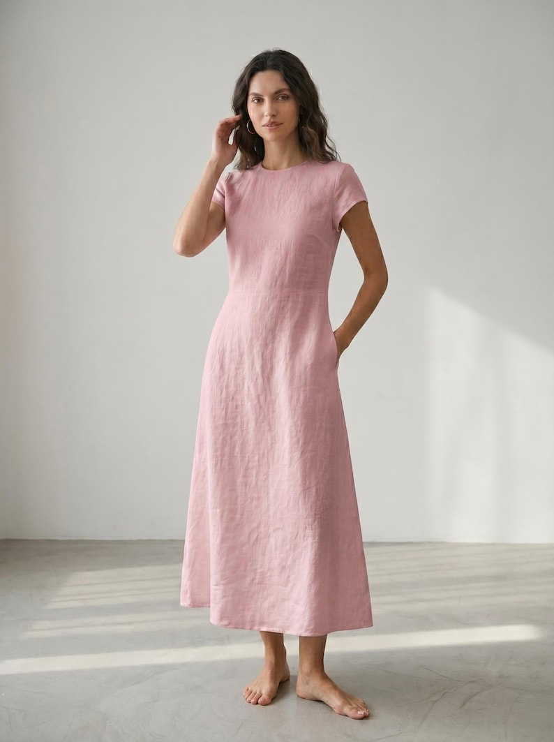 May include: A light pink midi dress with short sleeves and a round neckline. The dress has a fitted bodice and a flared skirt, with pockets on each side. The model is wearing the dress and standing in a room with natural light.