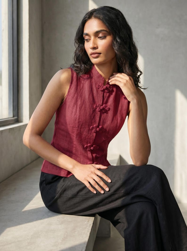 May include: A sleeveless burgundy top with a mandarin collar and decorative knot closures. The top is paired with black pants. The outfit is made of a lightweight fabric, suitable for warm weather. The style is reminiscent of traditional Asian designs.