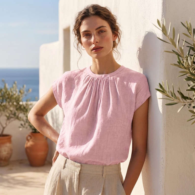 Peut inclure: Un haut en lin rose clair avec un col fronc&eacute; et des manches courtes. Le haut est associ&eacute; &agrave; un pantalon en lin beige. L'arri&egrave;re-plan pr&eacute;sente un mur blanc, des oliviers et une vue sur la mer.